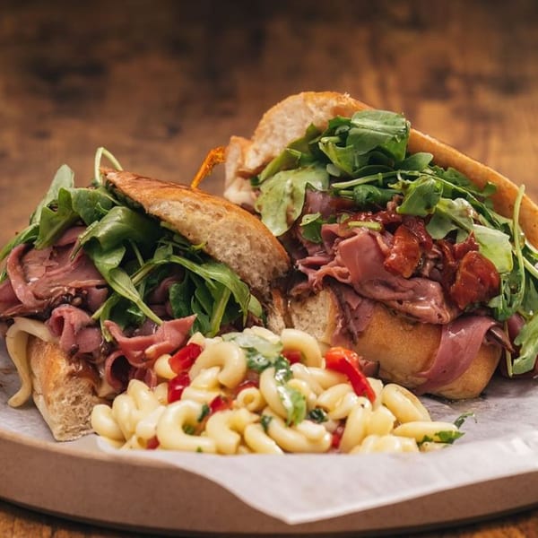 Two Italian beef sandwiches with arugula and a side of macaroni salad on a wooden table.